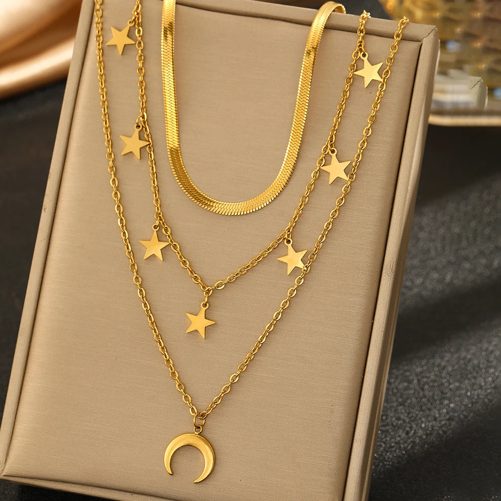 A gold-toned layered necklace set displayed on a beige jewelry box. The set includes a thick snake chain, two delicate chain layers with star charms, and a crescent moon pendant.